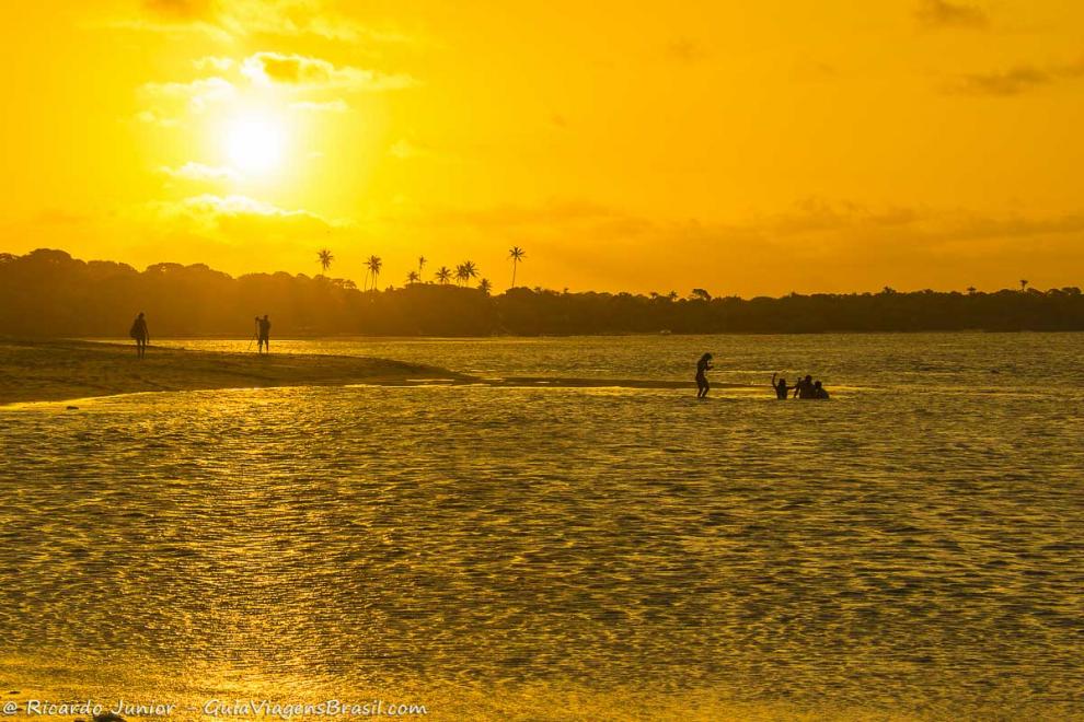 Imagem de turistas no mar e o sol se pondo ao fundo na Praia da Boca da Barra.